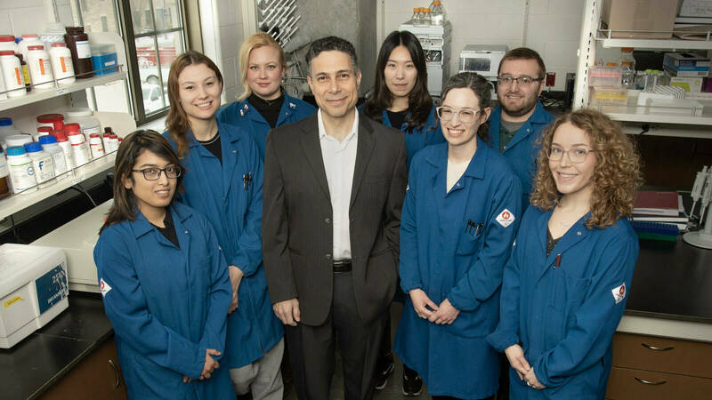 All lab members in 2023, smiling in blue lab coats, around advisor in black coat.