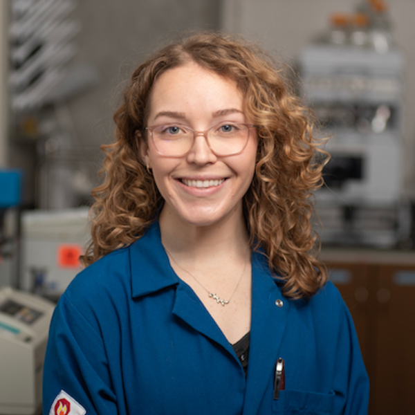 Smiling woman with curly auburn hair, wearing glasses and a blue lab coat.