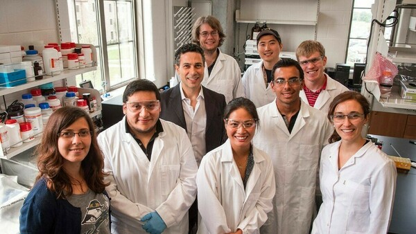 All lab members in 2016, smiling in white lab coats, around advisor in black coat.