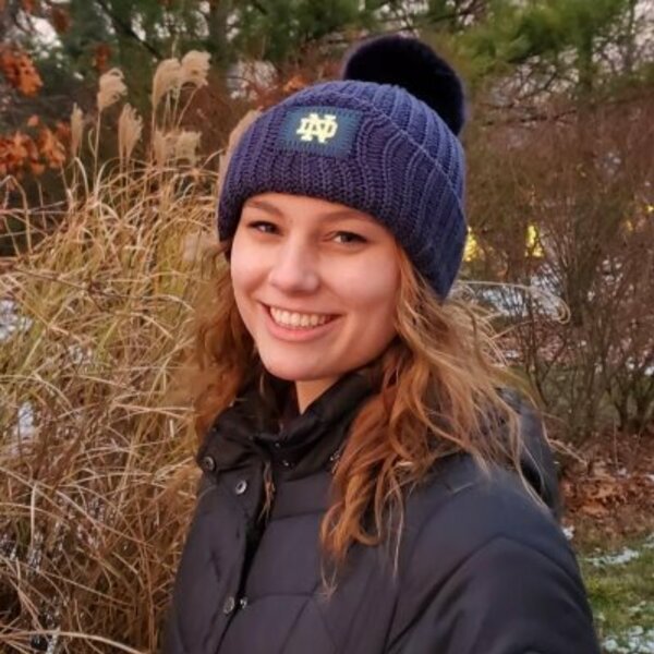 Smiling woman with curly auburn hair in Notre Dame beanie with Autumn background.